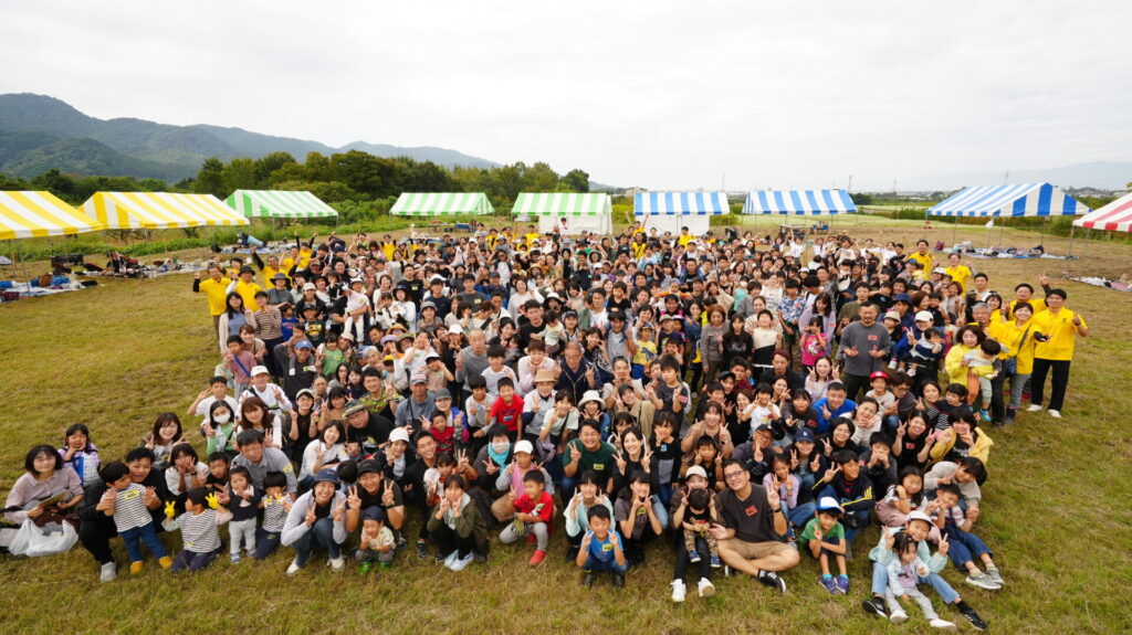 健康住宅のオーナー様感謝祭での集合写真。青空の下、広大な芝生広場に集まった数百組のご家族とスタッフの笑顔あふれる交流。