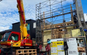 新築工事現場の足場とクレーン車。手前には「安全第一」の看板と健康住宅の施工事例パネルが並ぶ、整頓された建築現場。