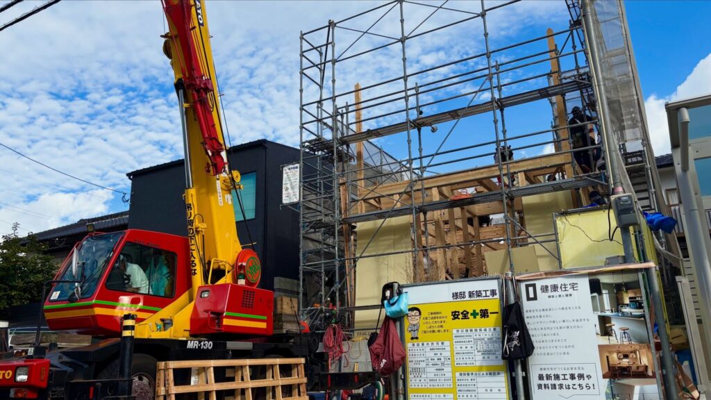 新築工事現場の足場とクレーン車。手前には「安全第一」の看板と健康住宅の施工事例パネルが並ぶ、整頓された建築現場。