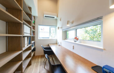 大容量の壁面本棚を備えた造作ワークスペース。横長のカウンターデスクと、手元を照らすブラケットライト、高窓から光が差し込む集中できる空間。