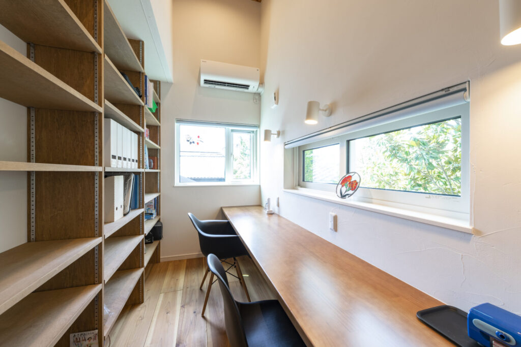 大容量の壁面本棚を備えた造作ワークスペース。横長のカウンターデスクと、手元を照らすブラケットライト、高窓から光が差し込む集中できる空間。